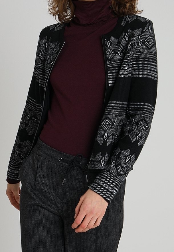 Personne portant un cardigan zippé à motif noir et gris par-dessus un col roulé bordeaux et un pantalon gris avec cordon de serrage.