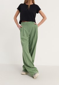 Zwart cropped t-shirt gecombineerd met hoge taille, geplooide groene wijde broek. Zacht materiaal, twee voorzakken en een knoopdetail.