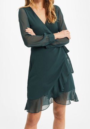 Žena v tmavě zelených zavinovacích šatech s výstřihem do V, průsvitnými dlouhými rukávy a volánkovým lemem, se zkříženýma rukama, stojí před bílým pozadím.