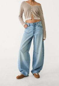 Vaaleansiniset leveälahkeiset farkut, joissa on etutaskut ja saumadetaljit, yhdistettynä lyhyeen beigen raidalliseen napilliseen neuletakkiin.