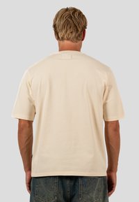 Beiges T-Shirt mit kurzen Ärmeln aus Baumwolle, mit Rundhalsausschnitt und einem kleinen Markenschild auf der Rückseite in der Nähe des Halsausschnitts.