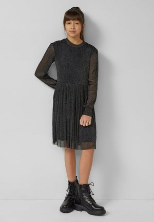 Schwarzes, langärmeliges Kleid mit funkelnder Textur, plissiertem Rock und transparenten Ärmeln. Kombiniert mit schwarzen Schnürstiefeln im Combat-Stil.