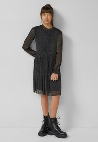 Zwarte jurk met lange mouwen, een sprankelende textuur, geplooide rok en doorschijnende mouwen. Gecombineerd met zwarte vetercombatlaarzen.