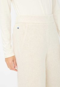 Cremefarbene Fleece-Hose mit glatter Textur, breitem Bund, Seitentaschen und einem kleinen, mehrfarbigen Logo-Akzent.