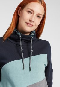 Sweatshirt color-blocké en bleu marine et teal avec un col montant haut, doté de cordons gris, en tissu doux et avec une coupe décontractée.