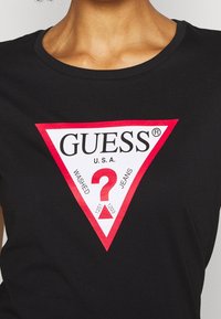 Sort T-shirt med et hvidt trekantet logo med rød kant, der står "GUESS U.S.A." over et stort rødt spørgsmålstegn og teksten "Washed Jeans".