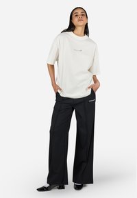 Camiseta blanca de gran tamaño con logotipo gráfico, combinada con pantalones anchos negros con texto blanco. El modelo está de pie con las manos en los bolsillos.