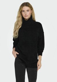 Maglione nero oversize con colletto alto, tessuto morbido e maniche lunghe. Abbinato a pantaloni neri brillanti.