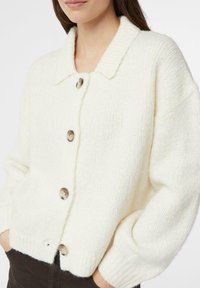 Biały dziergany kardigan z prążkowanym kołnierzem, dużymi guzikami i luźnym krojem z długimi rękawami. Widoczna fakturowana tkanina oraz boczne kieszenie.