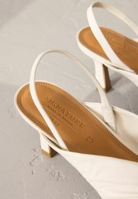 Weiße Slingback-Absätze mit quadratischer Zehenpartie, ausgestattet mit einem oberen Teil aus weichem Stoff und einer beigefarbenen Lederinnensohle, die mit "SIGNATURE MADE IN BRAZIL" geprägt ist.