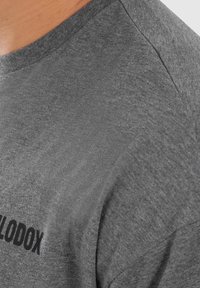 Camiseta de manga corta gris, hecha de una suave tela texturizada, con un logo negro impreso en la parte frontal. Cuello redondo simple y corte relajado.