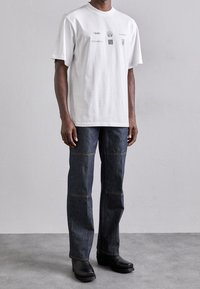Camiseta blanca de algodón con logotipos impresos, combinada con jeans de mezclilla oscura con costuras en contraste y botas negras hasta el tobillo.