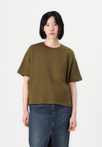 Olivgrünes, kurzärmliges Baumwoll-T-Shirt mit rundem Halsausschnitt und einer Brusttasche, kombiniert mit einem dunklen Jeansrock mit einem Schlitz vorne.