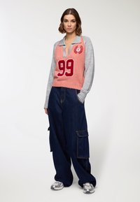 Maglione a blocchi di colore in rosa e grigio, con scollo a V profondo e logo con il numero "99". Pantaloni cargo in denim scuro dalla vestibilità ampia, con tasche laterali.