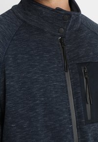 Giacca di un blu scuro con texture, colletto alto, due bottoni automatici neri e due tasche verticali con zip sul petto.