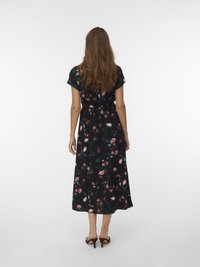 Γυναίκα με μακριά καστανά μαλλιά, φορώντας ένα μαύρο με λουλούδια μίντι φόρεμα και καφέ γόβες, που στέκεται με την πλάτη γυρισμένη απέναντι σε απλό φόντο.