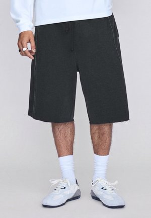 Homme portant un short gris foncé jusqu'aux genoux, des chaussettes blanches, des baskets blanches et une chemise blanche à manches longues, debout devant un fond uni.