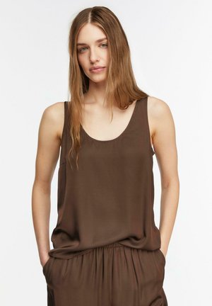 Jeune femme aux longs cheveux châtain clair portant un haut marron sans manches et un pantalon assorti, mains dans les poches, expression neutre.