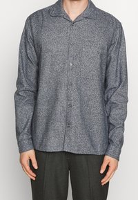 Mężczyzna w długim, zapinanym na guziki szarym teksturowanym koszuli, stojący z rękami swobodnie opuszczonymi po bokach, ubrany w ciemne, kraciaste spodnie.