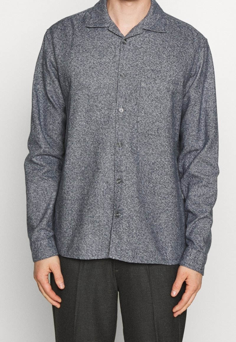 Mężczyzna w długim, zapinanym na guziki szarym teksturowanym koszuli, stojący z rękami swobodnie opuszczonymi po bokach, ubrany w ciemne, kraciaste spodnie.