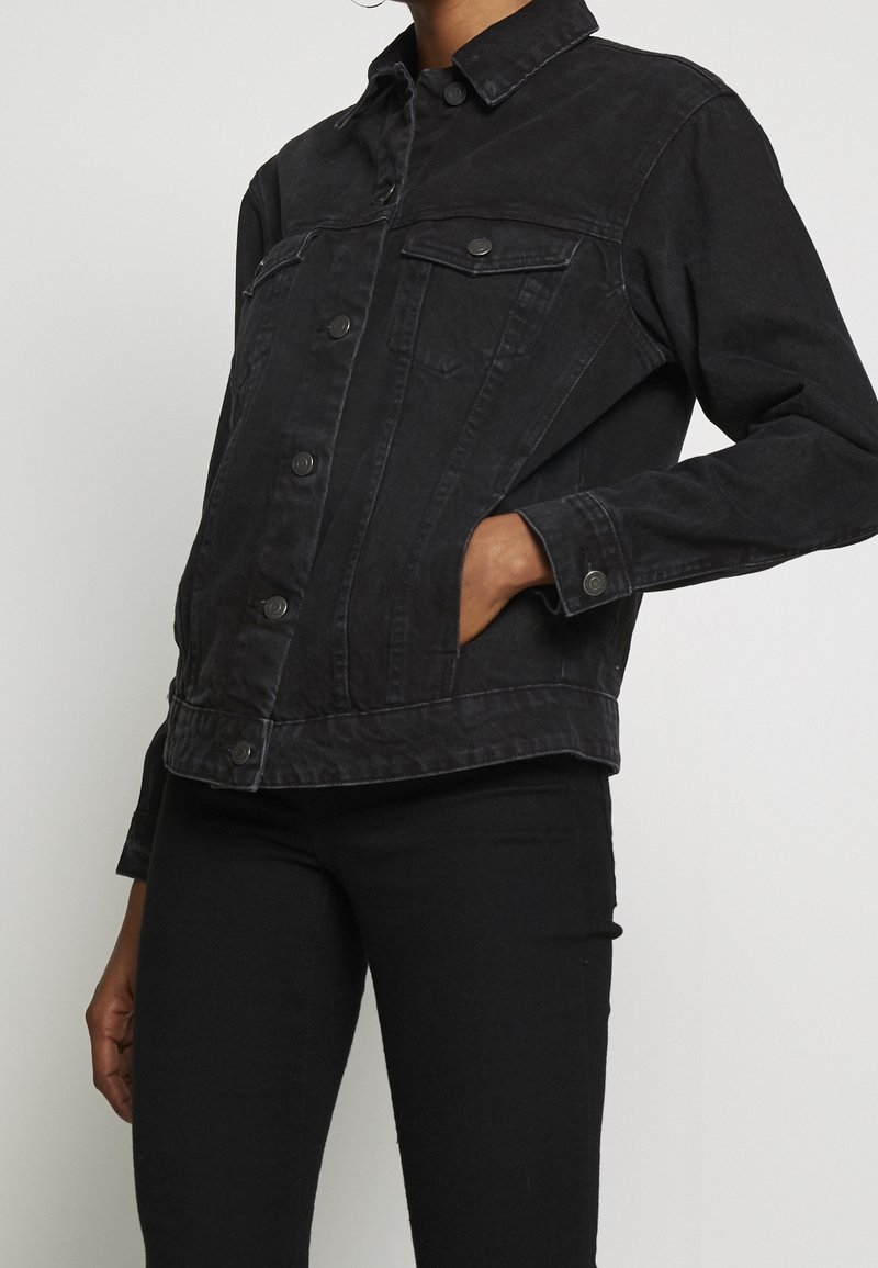 Personne portant une veste en jean noire avec des poches poitrine à boutons et des poches latérales, associée à un pantalon noir, sur un fond clair uni.