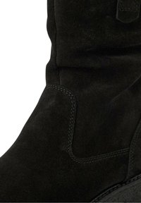 Gros plan sur une botte en daim noir montrant les coutures détaillées et le matériau doux et texturé sur le panneau latéral et la tige supérieure.