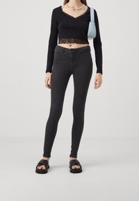 Top corto negro de manga larga con detalles de encaje, combinado con jeans ajustados negros de cintura alta. La modelo lleva sandalias negras y sostiene un bolso azul claro.