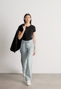 Ung kvinde går indendørs iført en sort t-shirt, lyseblå vidde-bukser, hvide sneakers og bærende en sort jakke over skulderen.