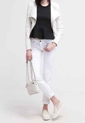 Femme portant un haut péplum noir, une veste et un pantalon blancs, tenant un sac à main blanc, avec des chaussures blanches sans lacets et les jambes croisées.