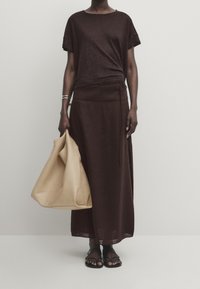 Maglietta marrone a maniche corte abbinata a una lunga gonna marrone fluente. Una borsa a tracolla in pelle beige completa l'outfit, indossata con sandali neri.