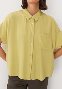 Camicia a maniche corte con bottoni, realizzata in un tessuto leggero di colore giallo chiaro, caratterizzata da una tasca sul petto e un orlo leggermente curvo.