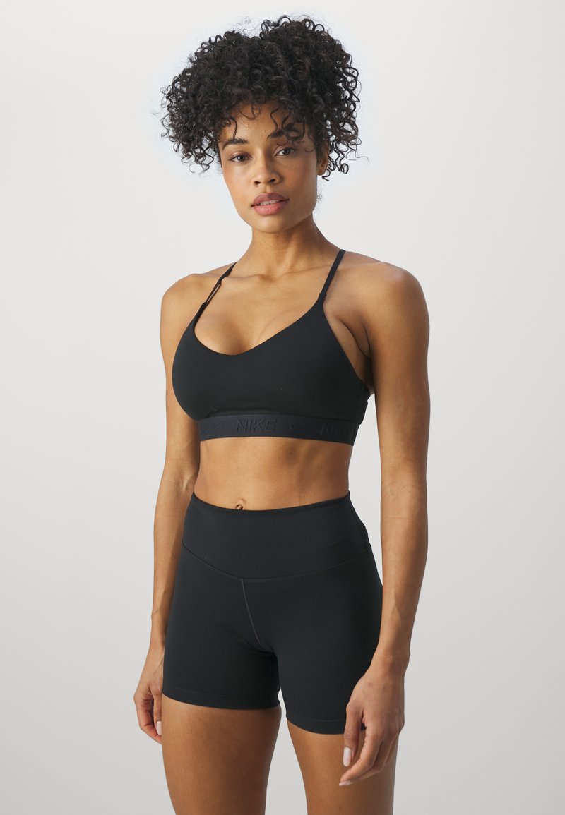 Sieviete ar cirtainiem matiem, valkājot melnu sporta krūšturi un augsta vidukļa melnas sporta šortus, stāv pret vienkāršu gaišu fonu.