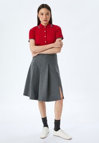 Polo shirt rosso a maglia a treccia con colletto bianco e dettagli con bottoni, abbinato a una gonna grigia plissettata con uno spacco laterale, indossato con sneakers bianche.