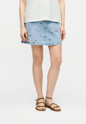 Frau trägt einen hellblauen Jeans-Minirock mit kleinen rosa-weißen Blumenstickereien, ein weißes Oberteil und braune Sandalen mit Riemchen.