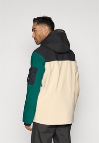 Casaco em bloco de cores nas tonalidades preto, verde e creme. Apresenta capuz, bolsos com fecho e uma textura de tecido adequada para uso ao ar livre.