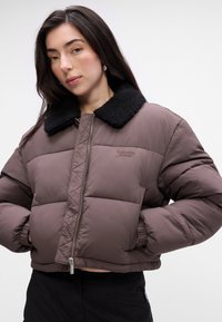 Brązowa kurtka puchowa z czarnym kołnierzem z owczej wełny, pikowana konstrukcja, zamek błyskawiczny z przodu, ściągacze przy mankietach oraz dyskretne logo.