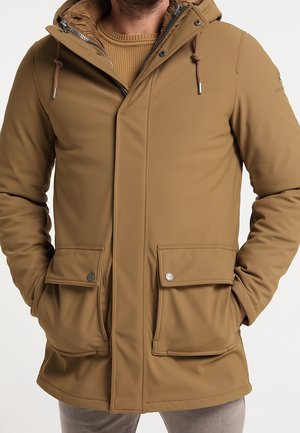Homme portant une veste d'hiver à capuche couleur sable avec une fermeture éclair à l'avant, des poches à rabat boutonnées et des détails à cordon de serrage, les mains dans les poches.