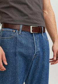 Ceinture en cuir marron avec une boucle en métal argenté, portée sur un jean en denim bleu ; design décontracté, cuir texturé et accents de couture visibles.