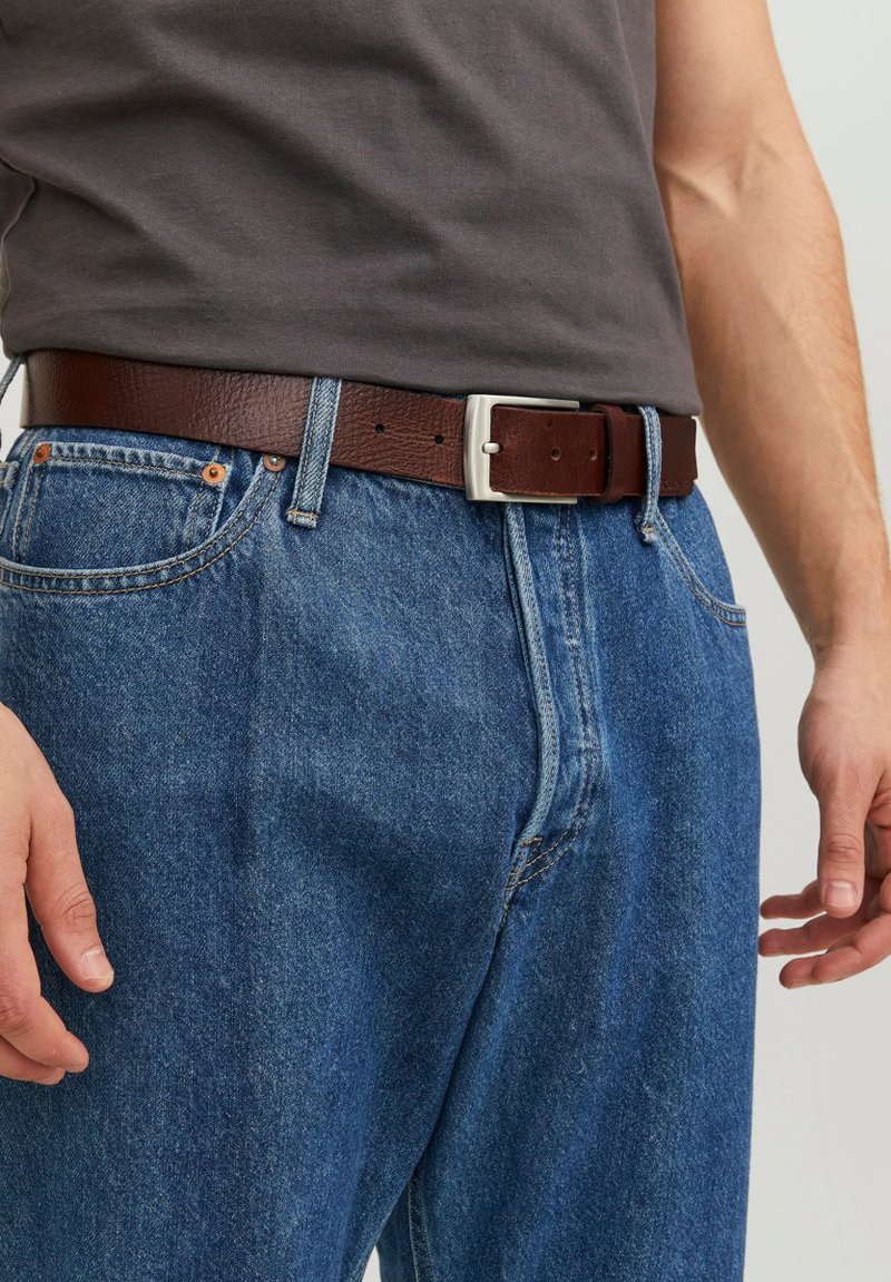 Ceinture en cuir marron avec une boucle en métal argenté, portée sur un jean en denim bleu ; design décontracté, cuir texturé et accents de couture visibles.
