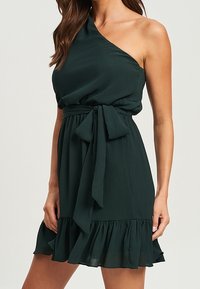 Ciemnozielona, jedno-ramienna sukienka z lekkiego materiału. Posiada przewiązany w talii pas i falbaniasty dół, co dodaje tekstury.