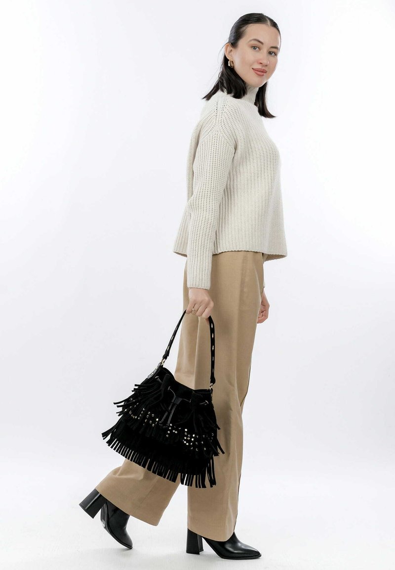 Borsa a tracolla in suede nero con dettagli di frange e borchie metalliche, tenuta da una persona in pantaloni beige e un maglione color crema, su uno sfondo bianco.