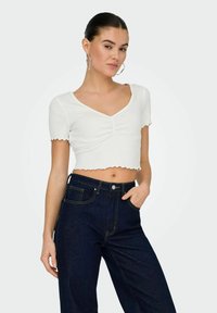 Witte cropped top met korte mouwen en een golvende zoom, gekenmerkt door een geraffineerde voorkant. Gepaarde met donkerblauwe high-waisted jeans.