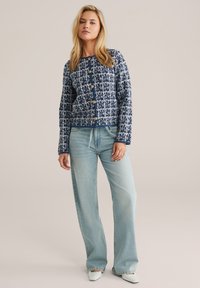 Navy and white patterned cardigan with gold buttons, paired with light blue wide-leg jeans and white pointed shoes. Smooth knit texture.
