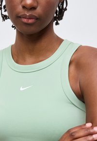 Κοντινό πλάνο ενός ατόμου που φορά ανοιχτό πράσινο μπλουζάκι χωρίς μανίκια της Nike, με πλεγμένα μαλλιά και σταυρωμένα χέρια.