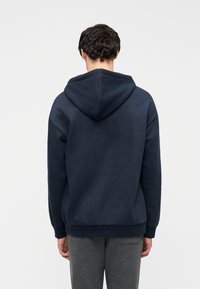 Μπλε καμπαρντίνα με μεγάλο, στρογγυλεμένο κουκούλι, μακριά μανίκια και ριμπ μανσέτες και στρίφωμα. Μαλακό ύφασμα με λεία υφή, χωρίς ορατά σχέδια.