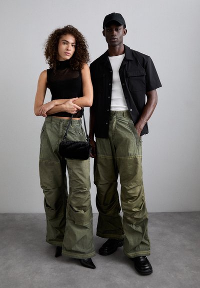 Schwarzes Mesh-Oberteil, olivgrüne Cargo-Hosen mit Taschen, schwarzes kurzärmliges Hemd, schwarze Mütze und strukturierte schwarze Handtasche. Glatter grauer Hintergrund.