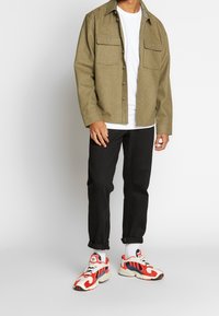 Olijfgroene shirtjack met twee borstzakken, gelaagd over een wit t-shirt, gecombineerd met zwarte broeken en rood-witte sneakers.