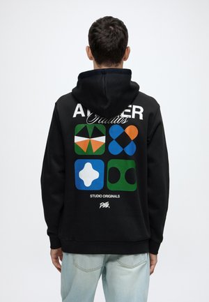 Mann trägt schwarzen Hoodie mit bunten geometrischen Formen und Text "STUDIO ORIGINALS" auf dem Rücken, dem Betrachter abgewandt.