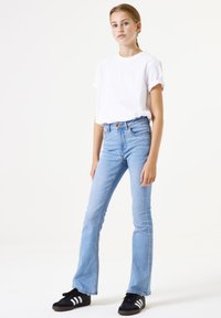 Weißes Kurzarm-T-Shirt, kombiniert mit hellblauen, ausgestellten Jeans und schwarzen Sneakers mit weißen Streifen. Schlichtes Design und lässiger Look.