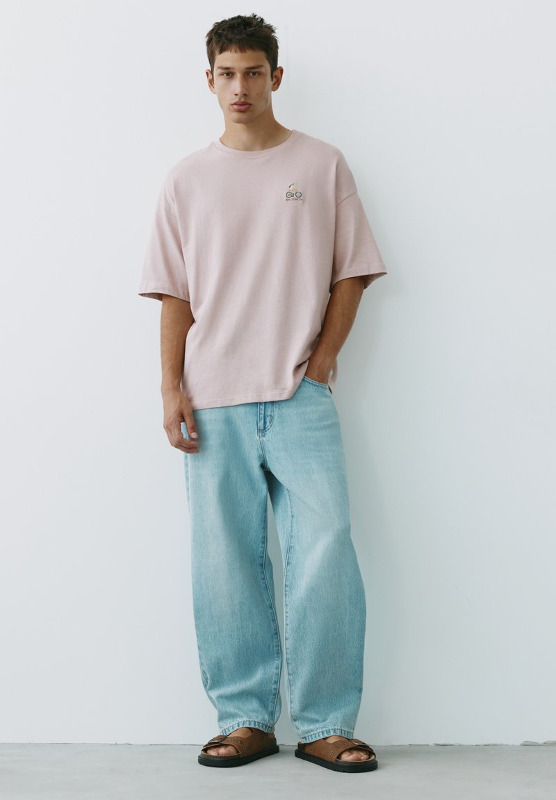 Jeune homme debout contre un mur blanc, portant un t-shirt oversize rose pâle, un jean bleu clair ample et des sandales marron.